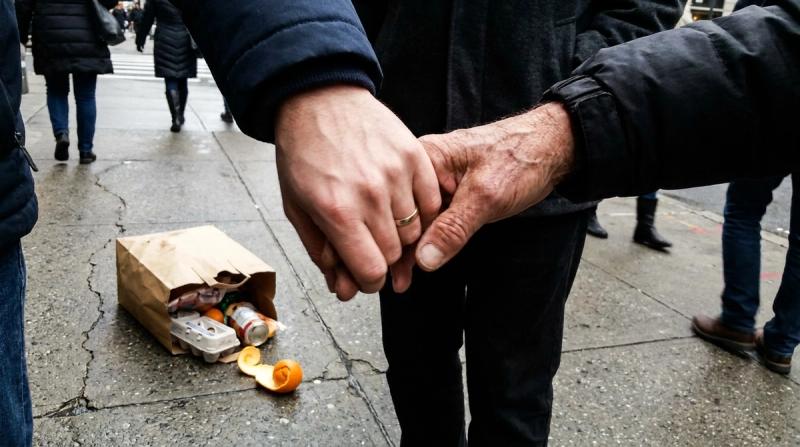 A young mans hand tightly holding an older mans hand on a city sidewalk, a split grocery bag nearby