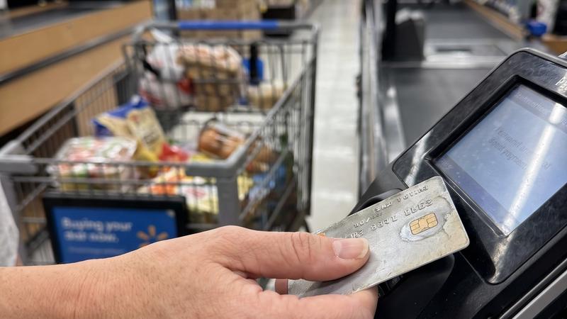 A woman's hand holding a bank card at a grocery store card reader, a full cart behind her