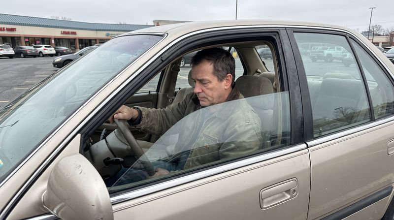 A man sitting alone in a parked older car in a store lot, seen from outside the window