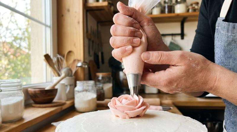 Hands piping a buttercream rose onto a cake with a pastry bag in a home bakery