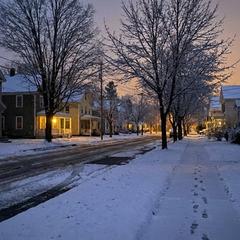 A snow-dusted small-town street at dusk with warm lights glowing in the windows of old wooden houses