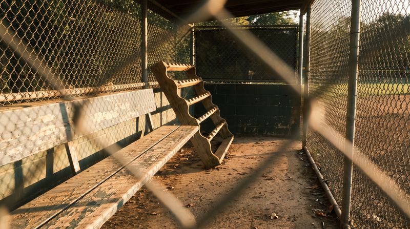 An empty youth baseball dugout with a chain-link fence and a bat rack