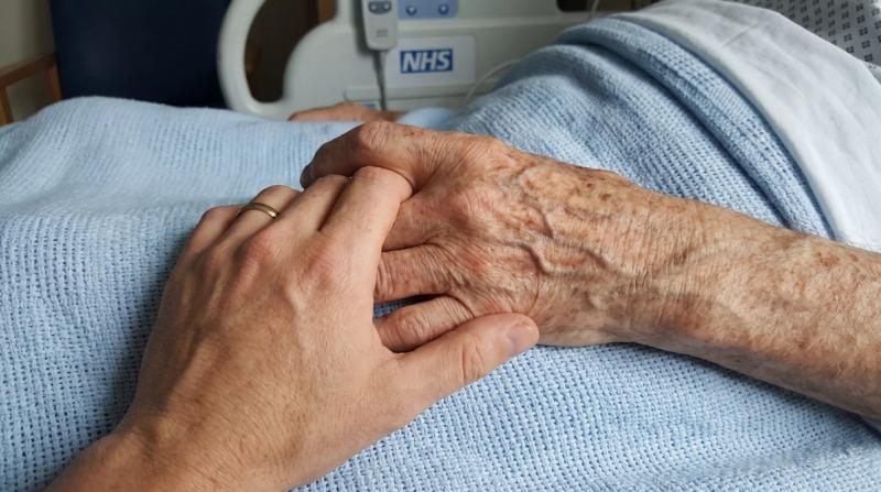 A younger hand holding the weathered hand of an elderly man on a hospital blanket