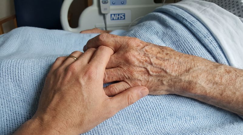 A younger hand holding the weathered hand of an elderly man on a hospital blanket