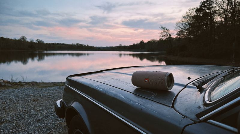 A small portable Bluetooth speaker sitting on the hood of a car beside a still reservoir at dusk