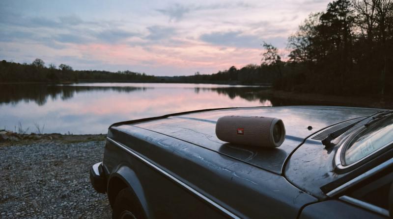 A small portable Bluetooth speaker sitting on the hood of a car beside a still reservoir at dusk