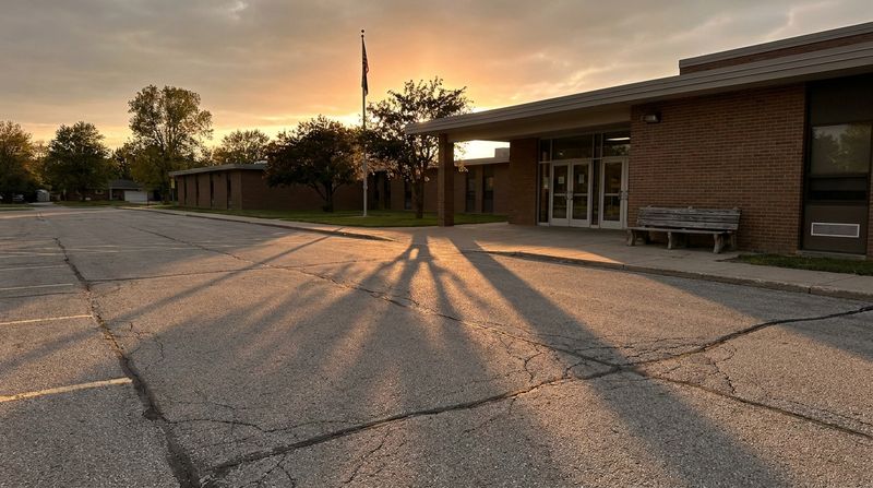 An empty school parking lot at dusk with long shadows and a single bench by the doors
