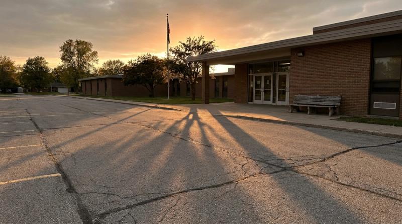An empty school parking lot at dusk with long shadows and a single bench by the doors