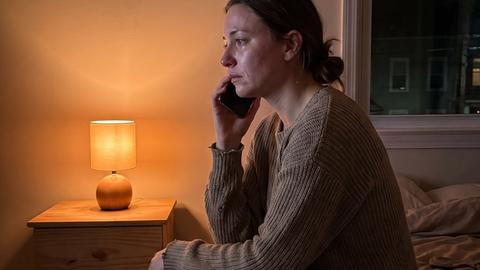 A woman sitting alone on the edge of a bed at night, holding a phone to her ear in the lamplight