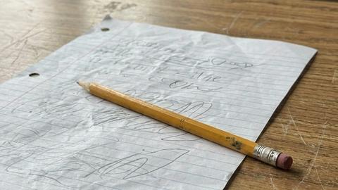 A worn yellow pencil resting on a crumpled sheet of lined paper on an old wooden school desk by a window