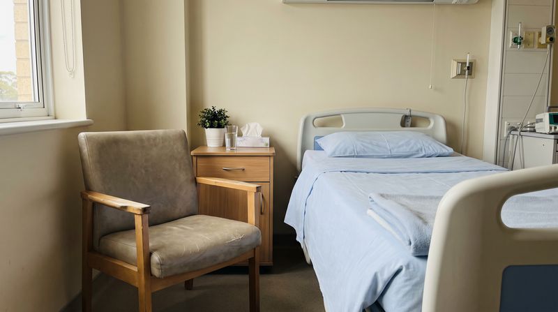 A quiet hospital room with an empty chair pulled up beside the bed in soft daylight