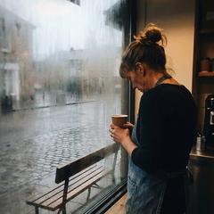 A weary widow in her fifties stands at her misty café window at dawn, holding a coffee cup and looking out at an empty bench, close to tears