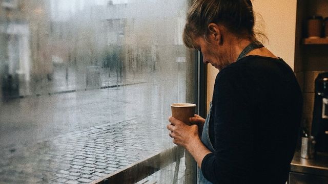 A weary widow in her fifties stands at her misty café window at dawn, holding a coffee cup and looking out at an empty bench, close to tears