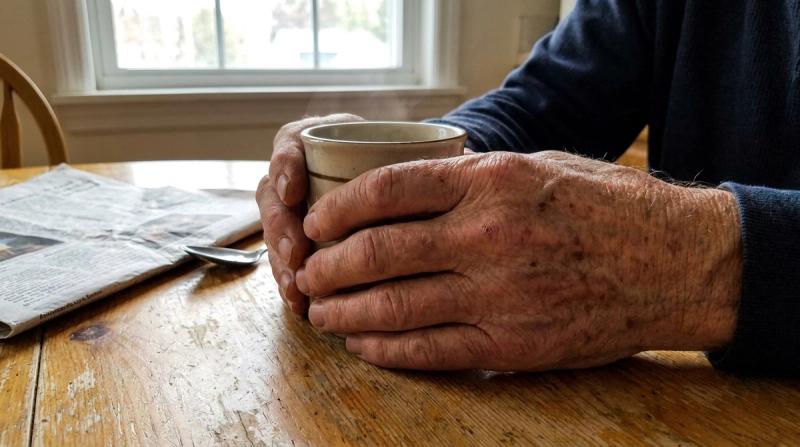 An older man's hands around a coffee mug at a small kitchen table in soft daylight
