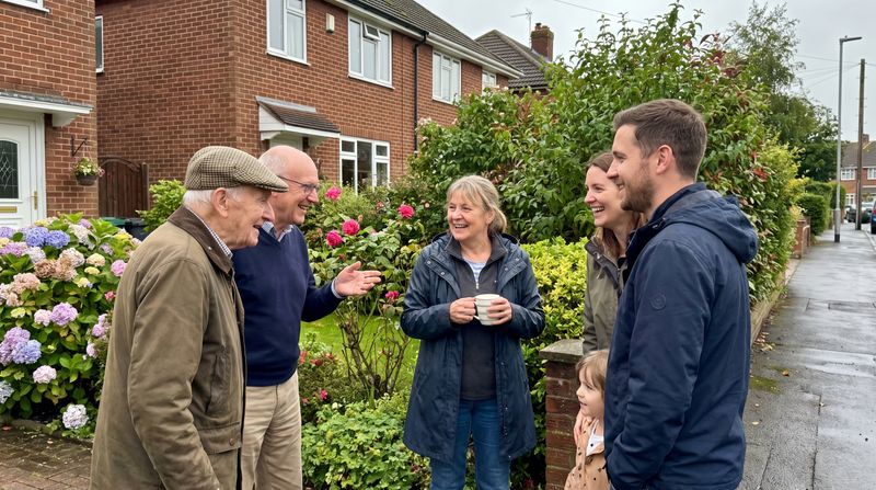 A group of neighbours of different ages standing together in a blooming front garden