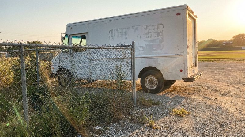 A delivery van parked on a gravel road behind a chain-link outfield fence