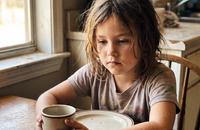 A young girl sitting alone at a kitchen table in warm morning light, seen from across the table