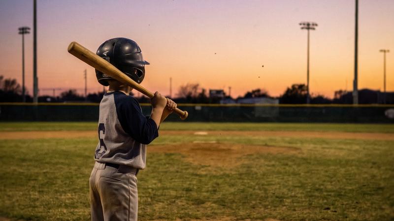 A small youth baseball player standing at home plate holding a bat, seen from behind at dusk