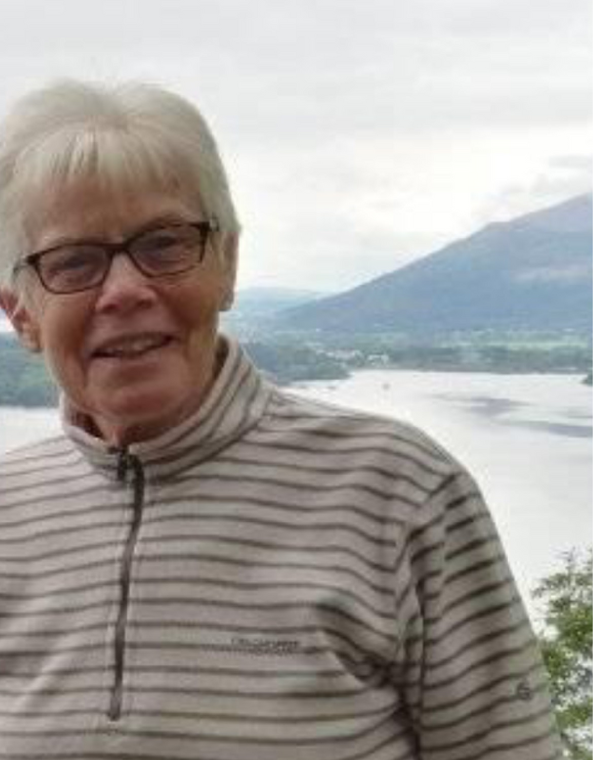 A woman with white hair and glasses smiling on a hillside, with a lake and a mountain behind her.
