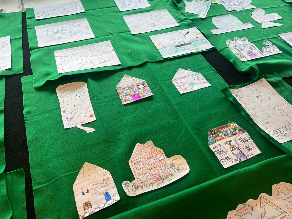 Coloured drawings of houses labelled with a variety of words and phrases such as "safe house" and "welcome". Some are coloured in, some are cut out of the paper. They are arranged in a rough grid on a green cloth.