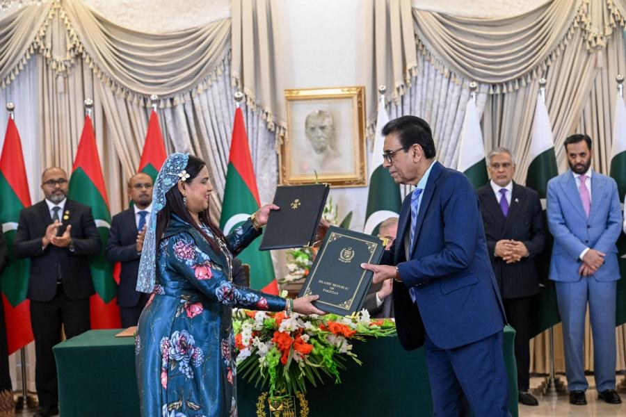 A Maldivian official in a blue patterned dress hands a folder to a Pakistani official in a blue suit.