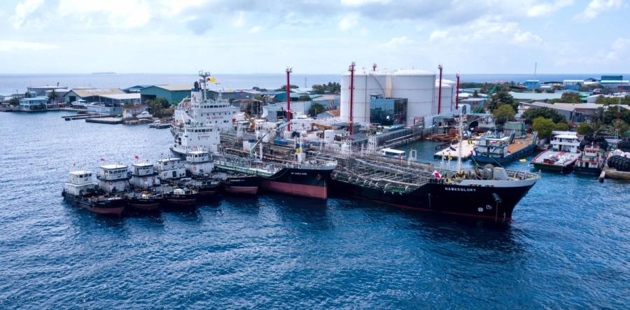Several tankers and barges moored side by side at a jetty in front of white storage tanks