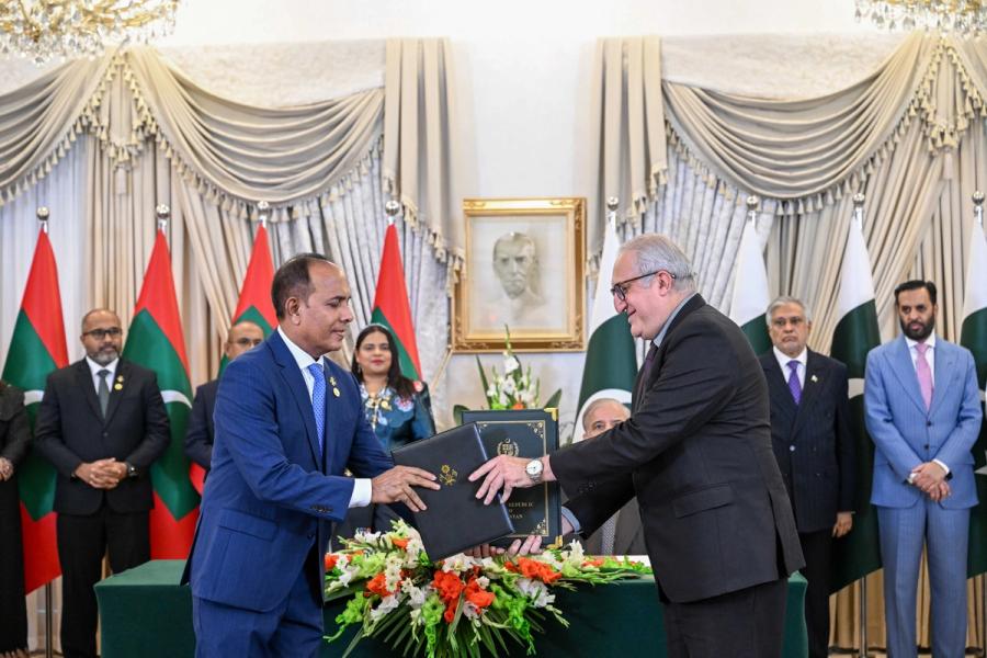 A Maldivian official in a blue suit exchanges a folder with a Pakistani official, with Pakistani officials applauding behind them.