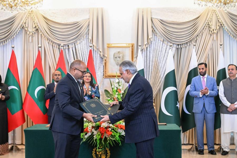 A Maldivian official and a Pakistani official exchange green folders across a table decorated with flowers.