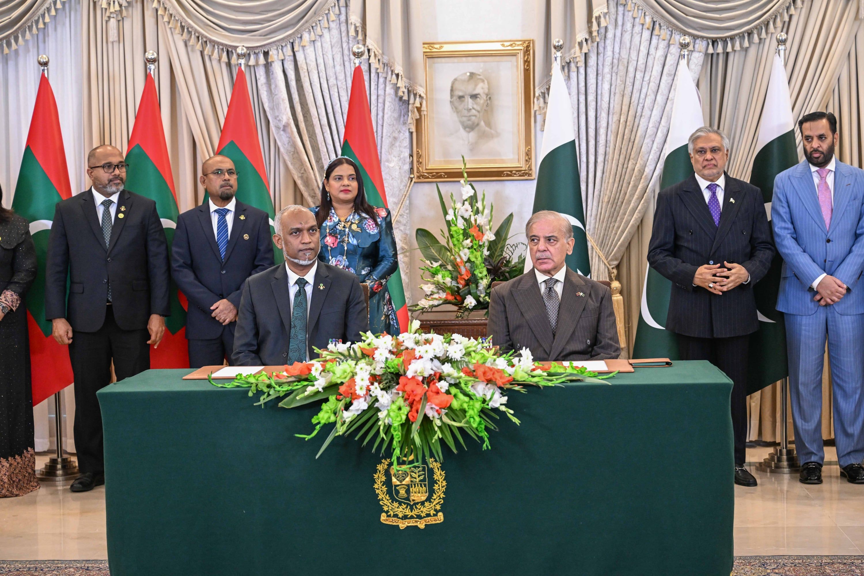 President Dr Mohamed Muizzu and Prime Minister Shehbaz Sharif seated side by side at a green signing table decorated with flowers, with Maldivian and Pakistani officials standing behind them in front of both countries’ flags.