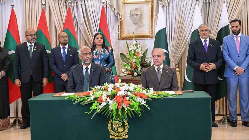 President Dr Mohamed Muizzu and Prime Minister Shehbaz Sharif seated side by side at a green signing table decorated with flowers, with Maldivian and Pakistani officials standing behind them in front of both countries’ flags.