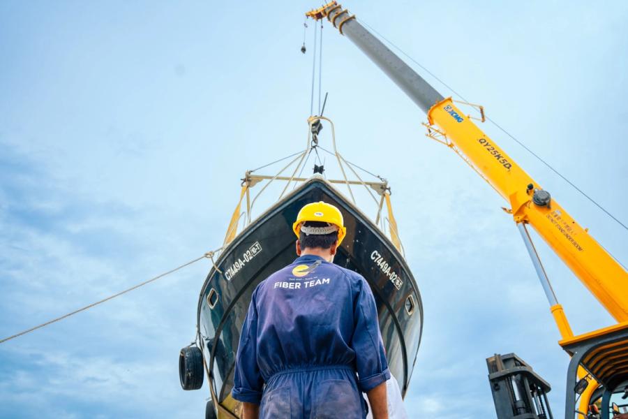 A worker in a hard hat and overalls marked Fiber Team watches a crane lift a boat hull