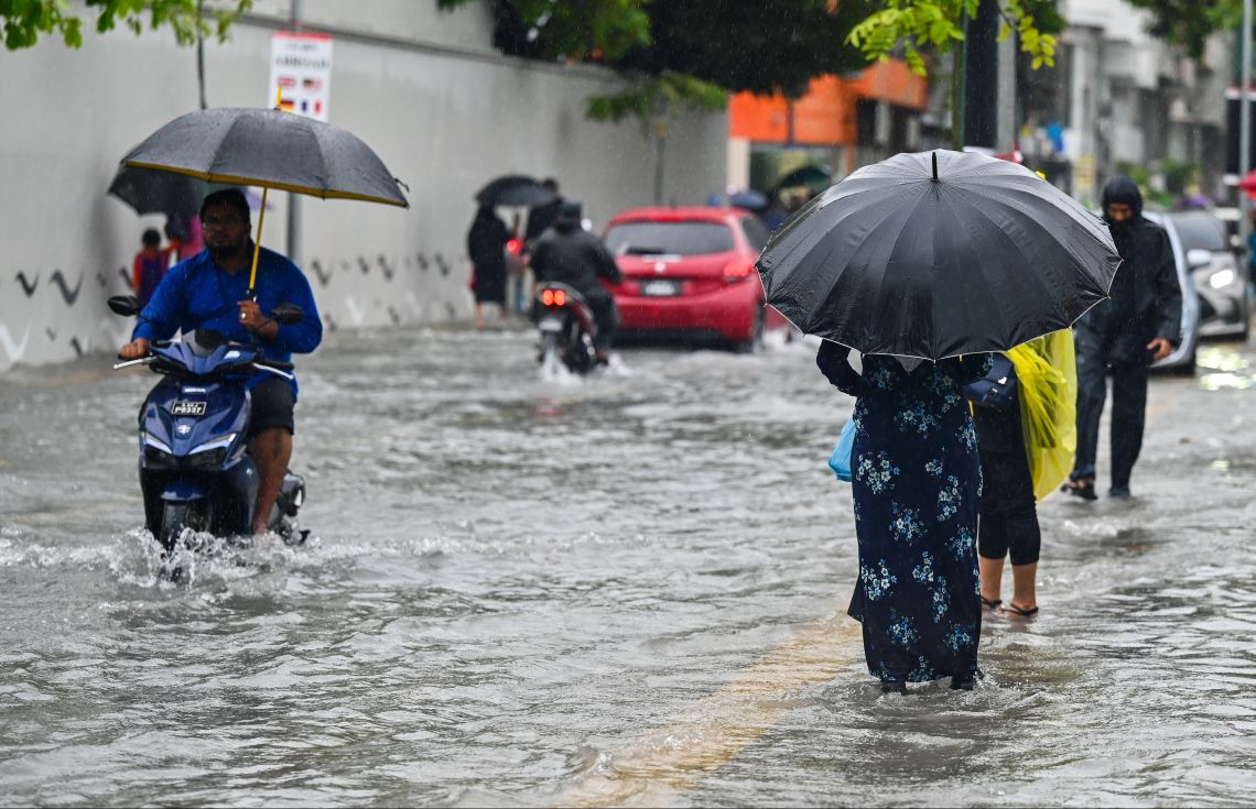 A man rides a scooter holding an umbrella and pedestrians wade through ankle-deep floodwater on a street in Malé during heavy rain.