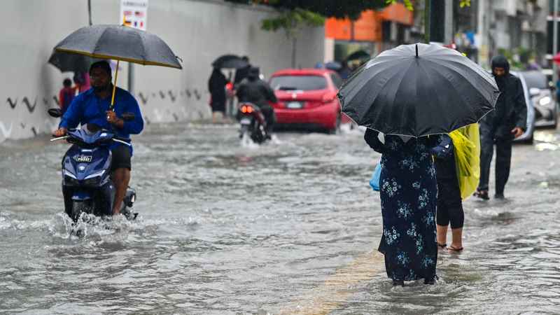 A man rides a scooter holding an umbrella and pedestrians wade through ankle-deep floodwater on a street in Malé during heavy rain.