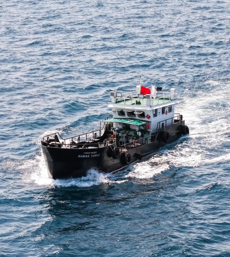 A small black-hulled fuel barge with a white wheelhouse under way at sea, seen from above