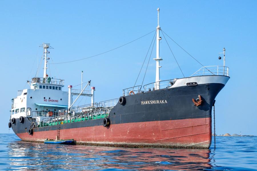 A small tanker with a black and red hull at anchor on calm water, the name Hawks Muraka on its bow