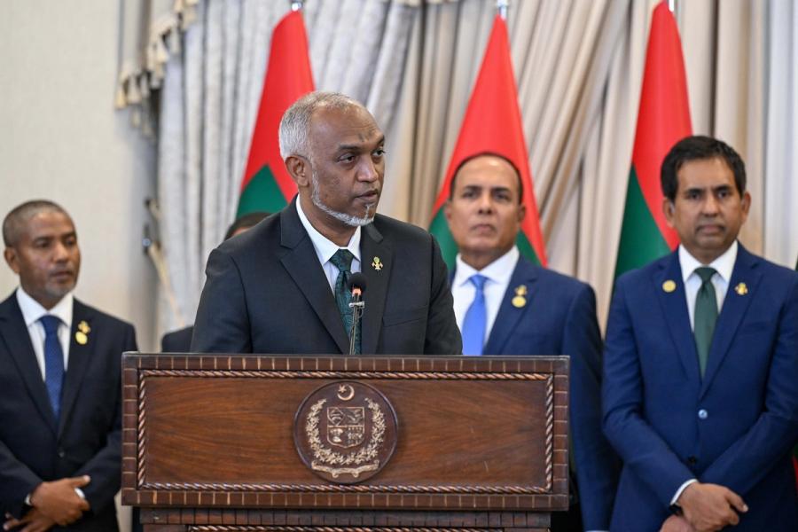 President Muizzu speaks at a carved wooden lectern in front of Maldivian flags, with members of his delegation behind him.