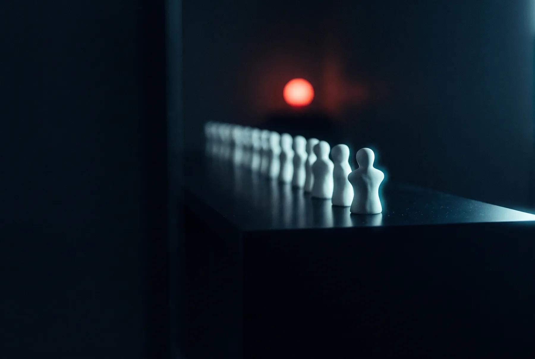 Hilera de figuras blancas idénticas que se pierde en la penumbra, con una al frente en foco y una luz roja al fondo