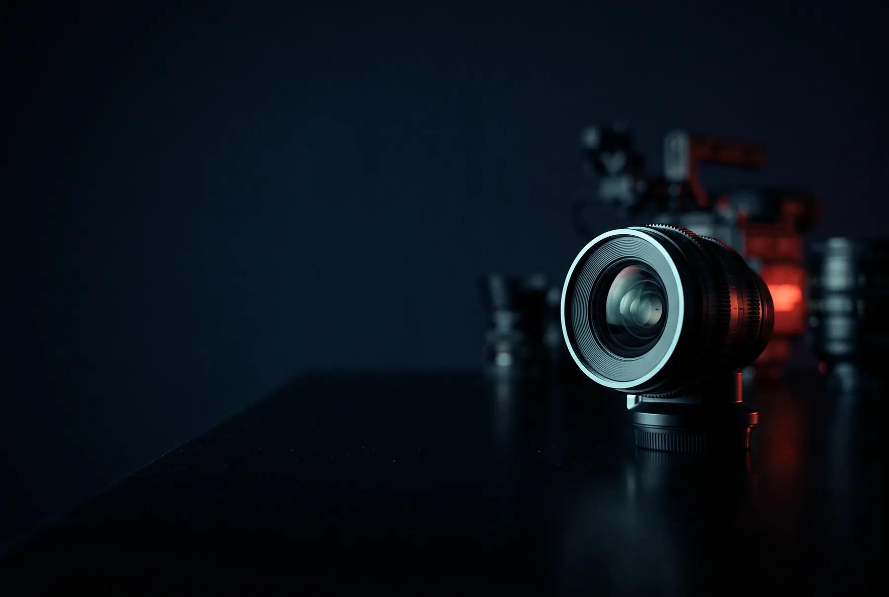 Lente de cine sobre una mesa oscura y reflectante, con equipo desenfocado y una luz roja al fondo