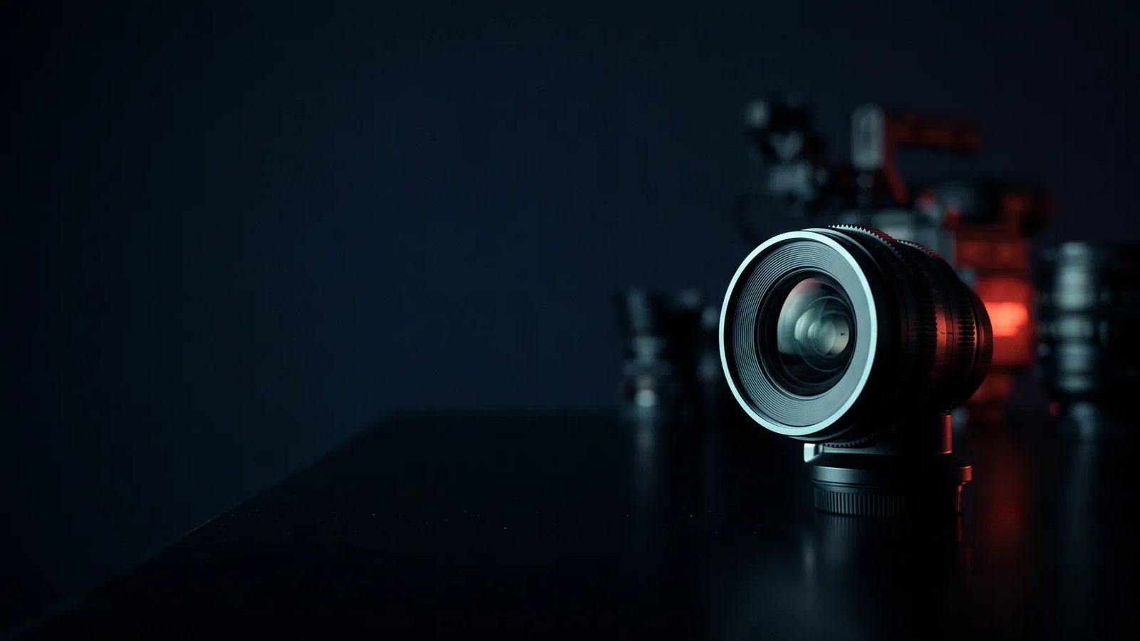 Lente de cine sobre una mesa oscura y reflectante, con equipo desenfocado y una luz roja al fondo
