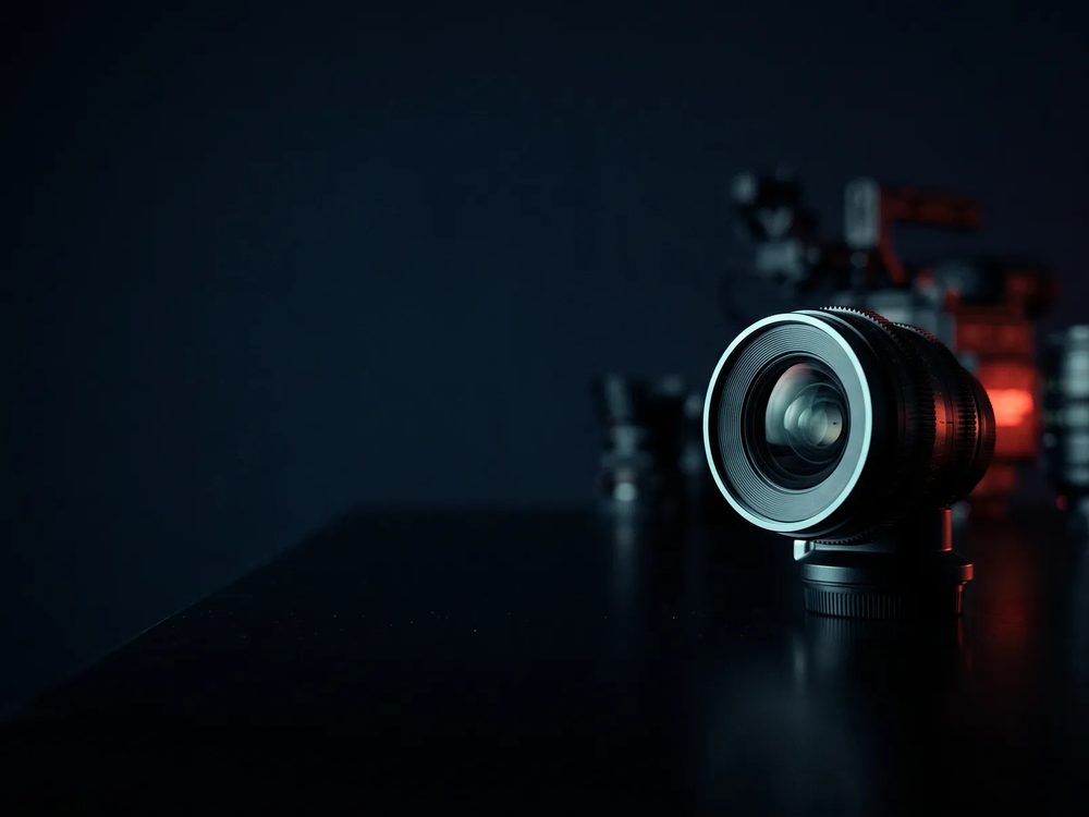 Lente de cine sobre una mesa oscura y reflectante, con equipo desenfocado y una luz roja al fondo