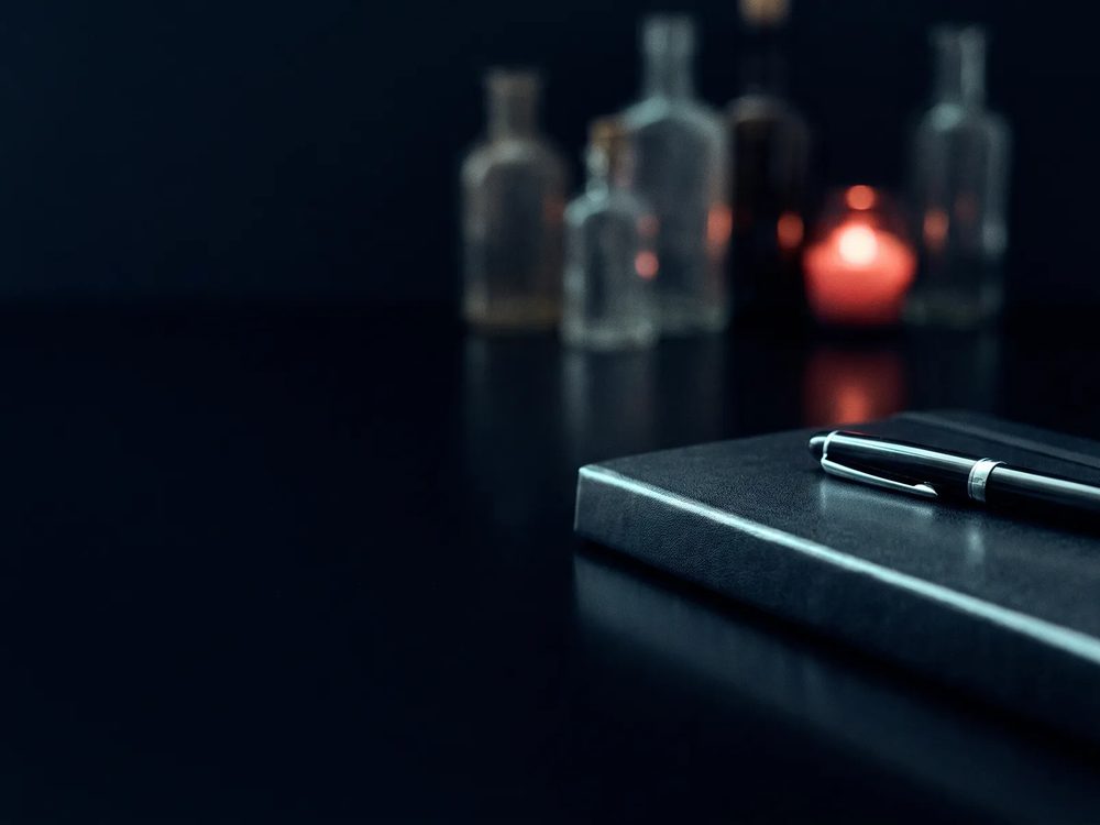 Cuaderno cerrado con una pluma encima sobre una mesa oscura y reflectante, con frascos desenfocados y una luz roja al fondo