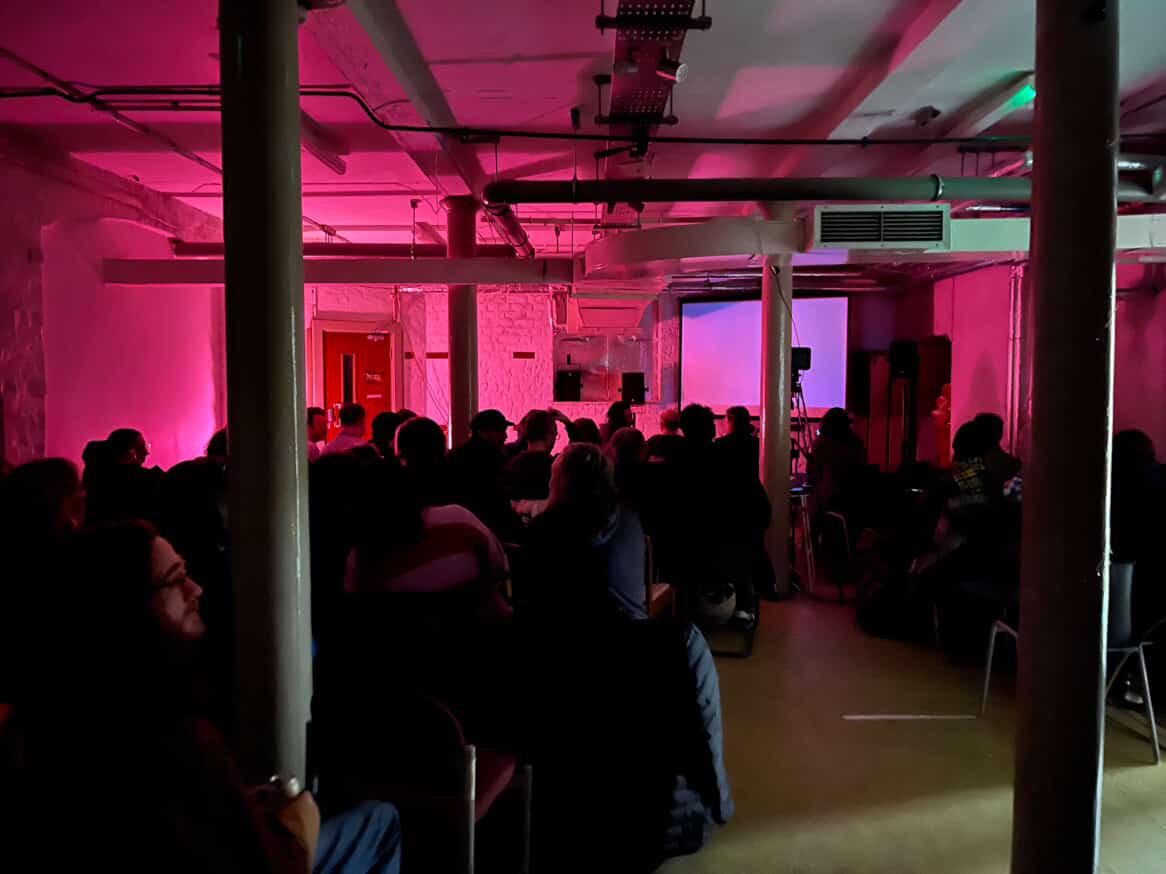 basement room full of people sat on chairs, looking towards a projection screen. the room is cast in pink light.