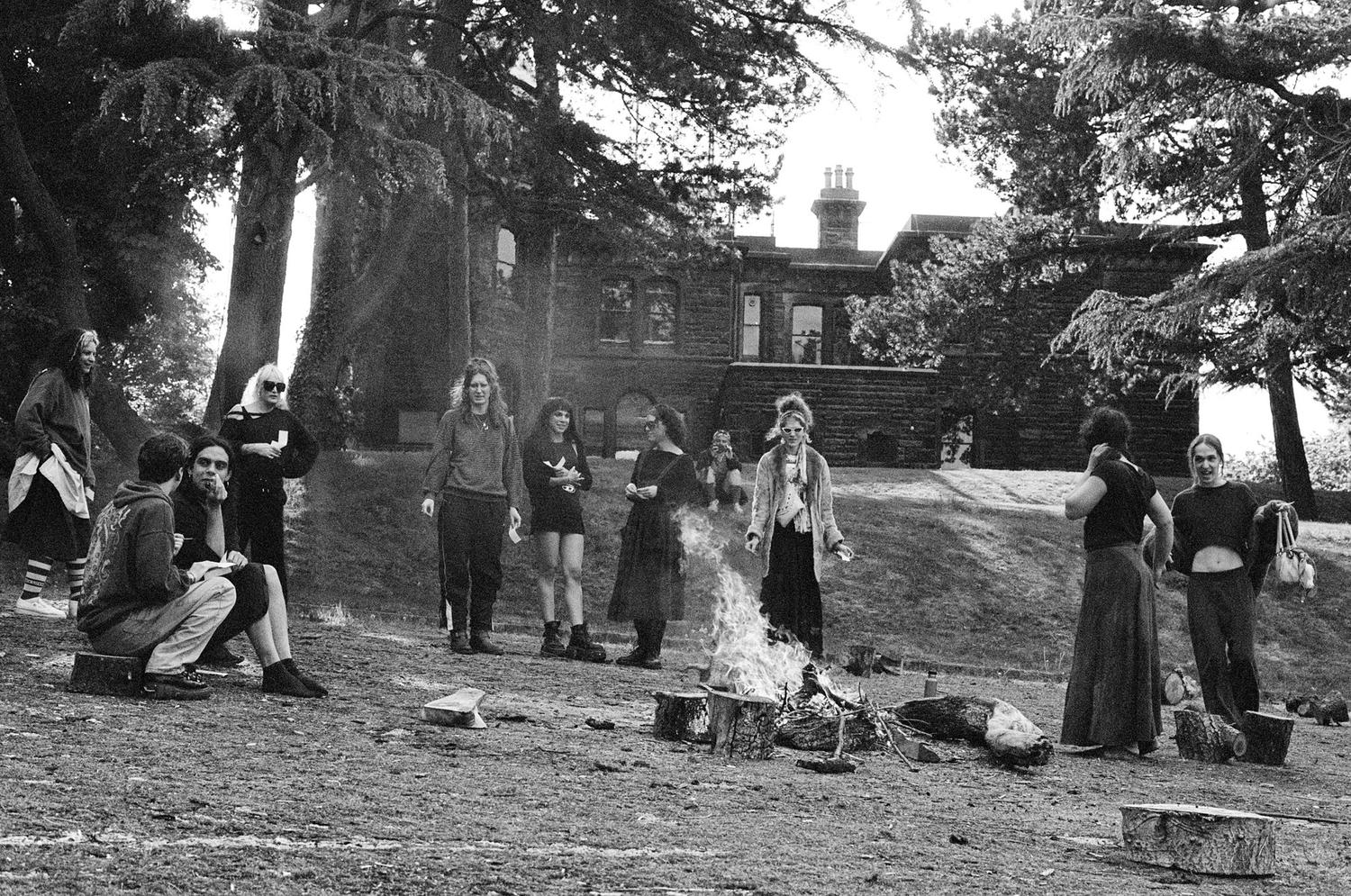 black and white photo of people stood around a fire, some sat on logs, bidston observatory and trees in the background