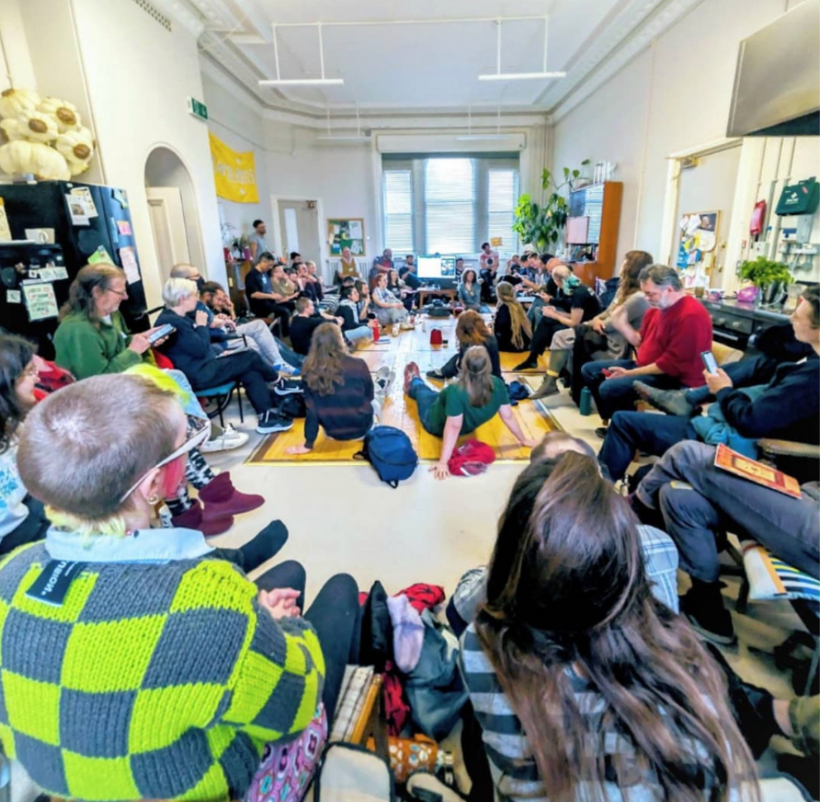 main kitchen space, full of people. most sat in a circle around the edge, and some sat on the floor, dotted with colourful mats and cushions.