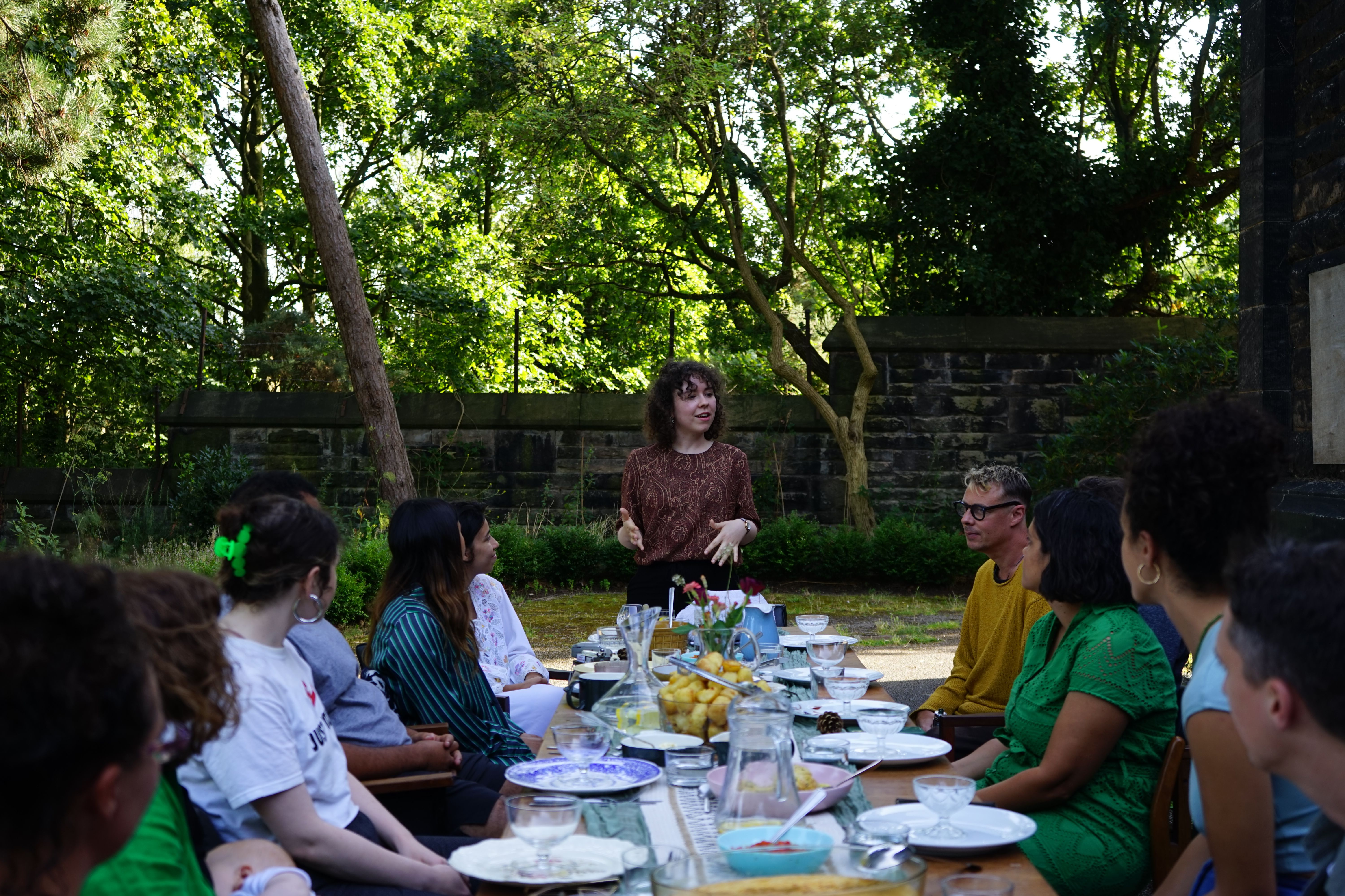 People gathered around a dinner table, outside in the sun, someone with a reddish brown top and curly hands stands at the head of the table, speaking