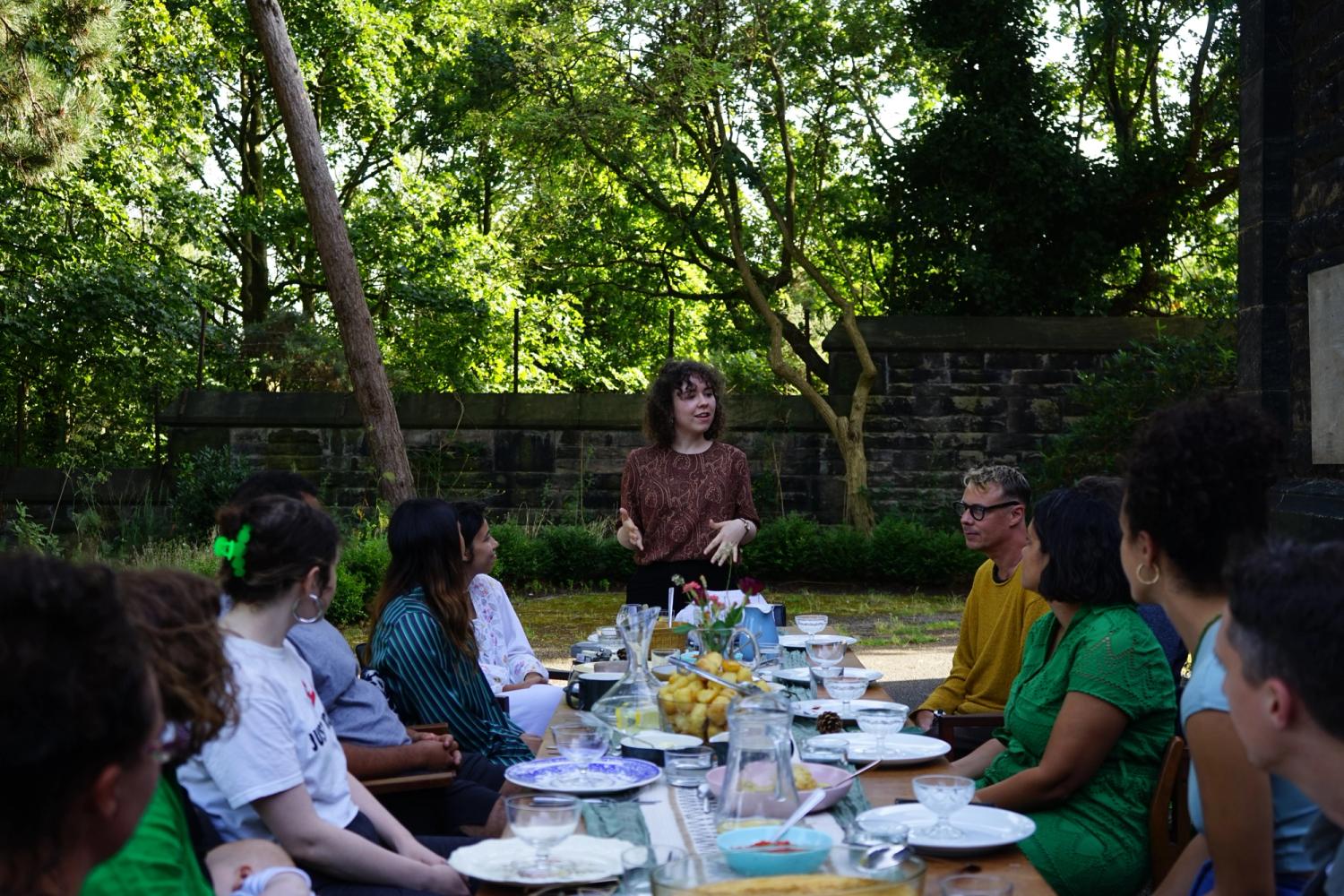 People gathered around a dinner table, outside in the sun, someone with a reddish brown top and curly hands stands at the head of the table, speaking