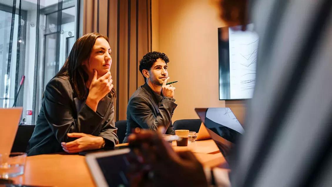 Two colleagues in a meeting room watching a presentation on screen