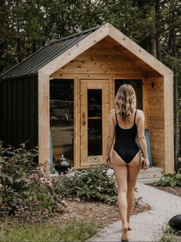 Woman walking toward a cedar cabin sauna in a backyard garden