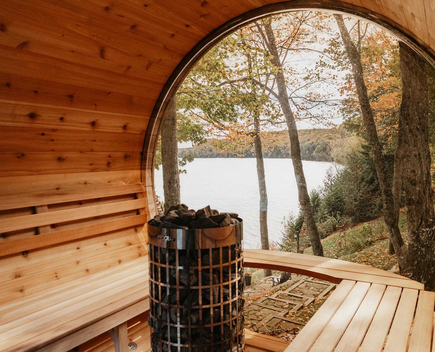 Cedar barrel sauna interior with a heater stacked with rocks, looking out a round porthole window onto an autumn lake