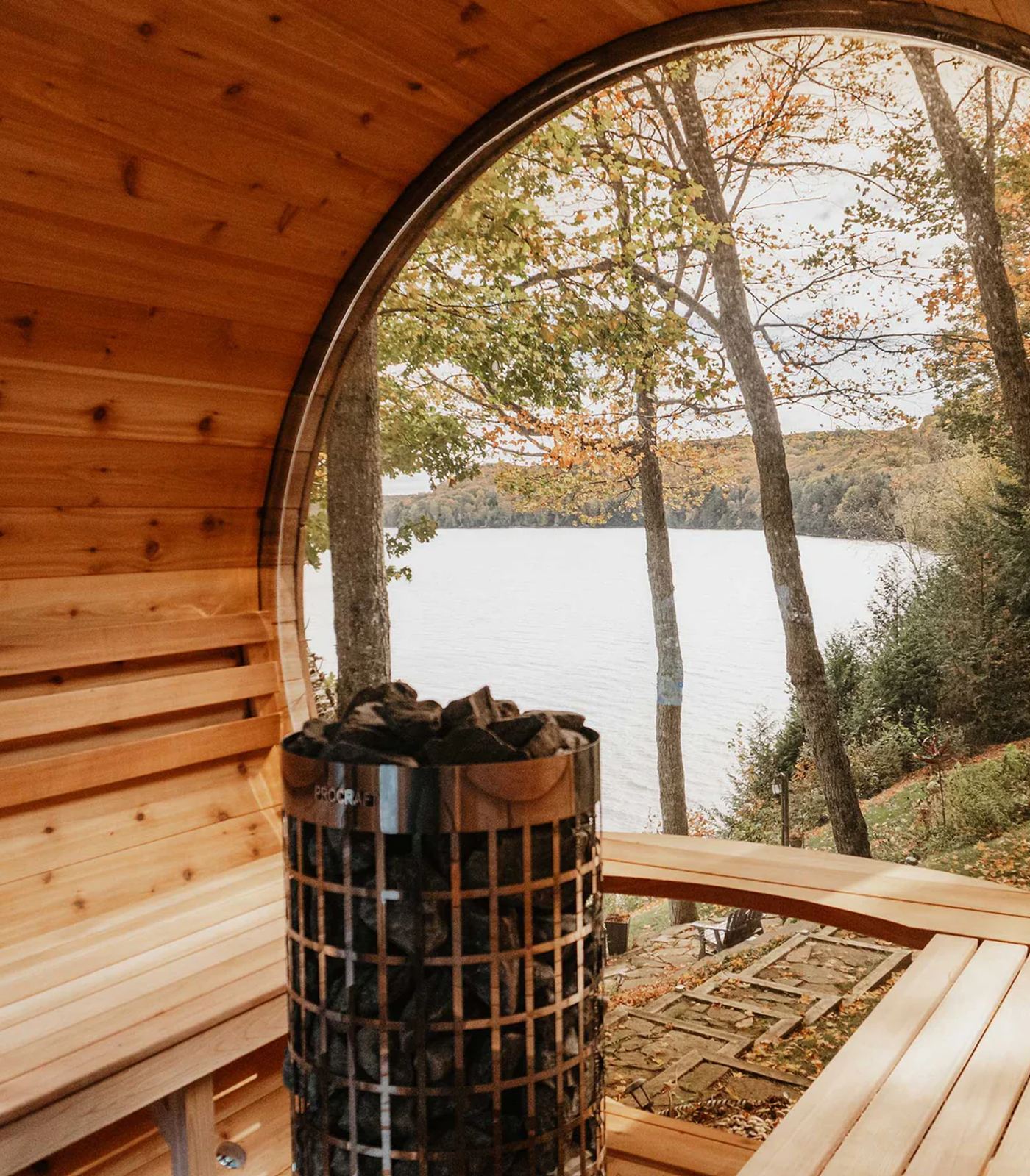 Cedar barrel sauna interior with a heater stacked with rocks, looking out a round porthole window onto an autumn lake